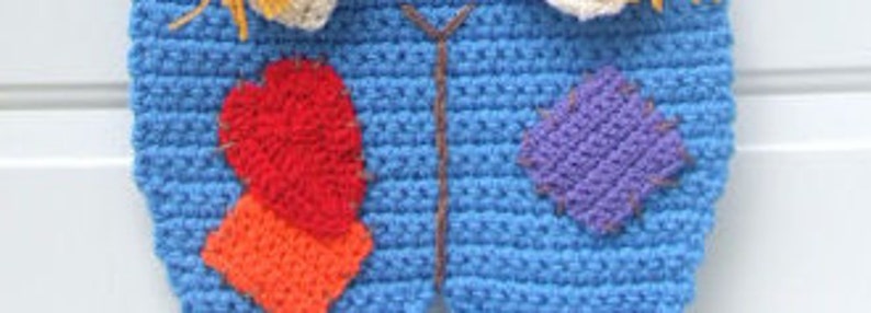 May include: A blue crocheted scarecrow costume with red, orange, and purple patches. The scarecrow has a straw hat and a brown crocheted belt.