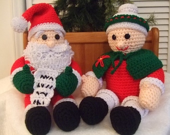 Puede incluir: Un Papá Noel y un duende de crochet. El Papá Noel lleva un gorro rojo, una barba blanca y guantes verdes. El duende lleva un gorro rojo y verde, un vestido rojo y una capa verde.
