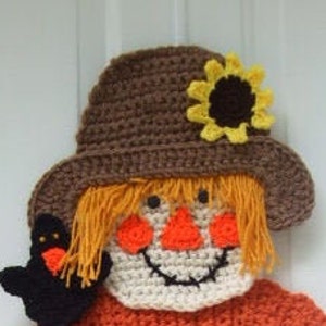 May include: Crocheted scarecrow head with a brown hat, orange hair, a white face, and a black bird. The scarecrow is wearing a brown hat with a yellow sunflower on top. The scarecrow has a black bird on its left side.