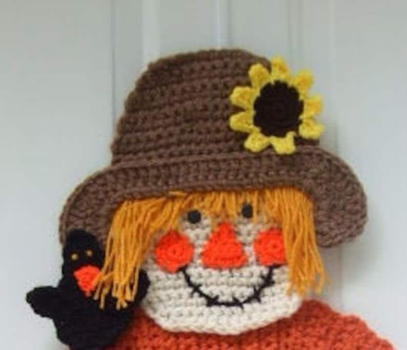 May include: Crocheted scarecrow head with a brown hat, orange hair, a white face, and a black bird. The scarecrow is wearing a brown hat with a yellow sunflower on top. The scarecrow has a black bird on its left side.