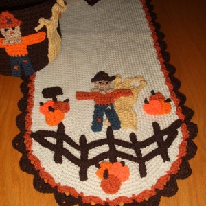 May include: A crocheted table runner with a brown border and a white center. The runner features a brown fence, orange pumpkins, and two brown and orange scarecrows.