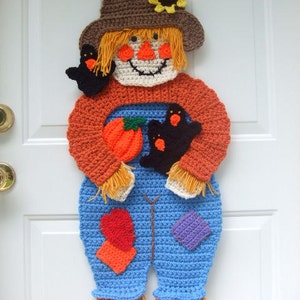May include: A crocheted scarecrow door hanger with a brown hat, orange shirt, blue overalls, and a pumpkin. The scarecrow has a yellow sunflower on its hat and two black birds in its hands.