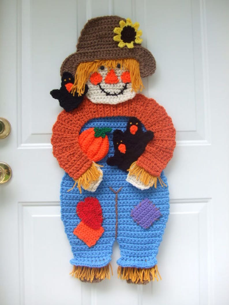 May include: A crocheted scarecrow door hanger with a brown hat, orange shirt, blue overalls, and a pumpkin. The scarecrow has a yellow sunflower on its hat and two black birds in its hands.