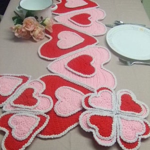 May include: A collection of handmade crochet heart-shaped placemats and coasters in shades of red and pink, with white trim. The placemats are arranged on a beige tablecloth, alongside a white plate, silverware, and a small bouquet of pink roses.