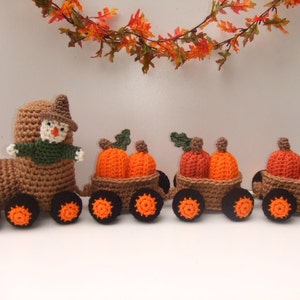 Puede incluir: Un tren de crochet con un conductor espantapájaros y tres vagones llenos de calabazas. El tren es marrón con ruedas naranjas y las calabazas son naranjas con tallos verdes.