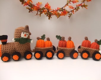 May include: A crocheted train with a scarecrow conductor and three pumpkin-filled carriages. The train is brown with orange wheels and the pumpkins are orange with green stems.