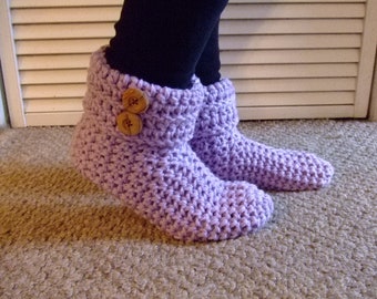May include: A pair of light purple crochet slippers with two brown wooden buttons on the side. The slippers are thick and cozy, perfect for indoor wear. The slippers are shown on a carpeted surface.