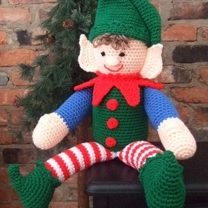 Può includere: Un pupazzo di elfo all'uncinetto con un cappello verde, pantaloni a righe rosse e bianche e scarpe verdi. L'elfo è seduto su una superficie di legno.