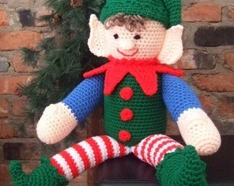 May include: A crocheted elf doll with a green hat, red and white striped pants, and green shoes. The elf is sitting on a wooden surface.