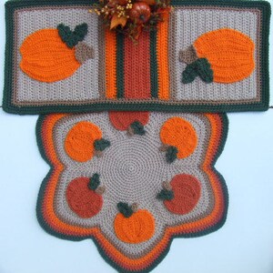 May include: A crocheted pumpkin-themed rug set with a runner and a round rug. The runner has a brown, orange, and green striped design with a pumpkin on each end. The round rug has a brown center with six orange pumpkins around the edge.