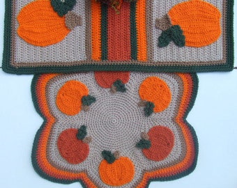 May include: A crocheted pumpkin-themed rug set with a runner and a round rug. The runner has a brown, orange, and green striped design with a pumpkin on each end. The round rug has a brown centre with six orange pumpkins around the edge.