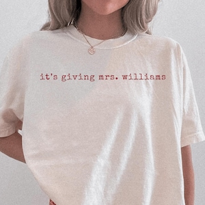 K&ouml;nnte beinhalten: Ein elfenbeinfarbenes Kurzarm-T-Shirt mit dem roten Schriftzug "it's giving mrs. williams" auf der Brust. Das Kleidungsst&uuml;ck hat einen Rundhalsausschnitt und eine lockere, weit geschnittene Passform, kombiniert mit einer rot-wei&szlig; karierten Shorts.
