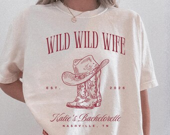 Puede incluir: Una camiseta color marfil con un estampado gráfico rojo con el texto WILD WILD WIFE, EST. 2026, Katie's Bachelorette y NASHVILLE, TN. El diseño incluye una ilustración de líneas de un sombrero de vaquero sobre un par de botas vaqueras.
