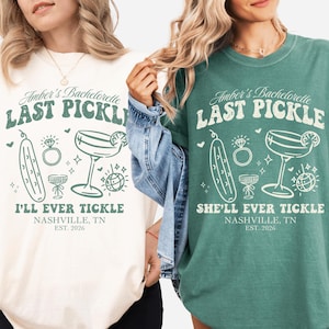 May include: Two t-shirts, one cream and one teal, featuring the text "Amber's Bachelorette LAST PICKLE" with illustrations of a pickle, ring, cocktail, and disco ball. The cream shirt reads "I'LL EVER TICKLE" and the teal shirt reads "SHE'LL EVER TICKLE".