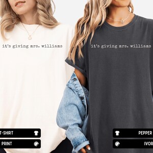 K&ouml;nnte beinhalten: Zwei kurz&auml;rmelige T-Shirts mit Rundhalsausschnitt, eines in Elfenbein und eines in Pfeffergrau, jeweils mit dem schwarzen Schriftzug 'it's giving mrs. williams' in Schreibmaschinen-Optik auf der Brust, in einer lockeren Passform.