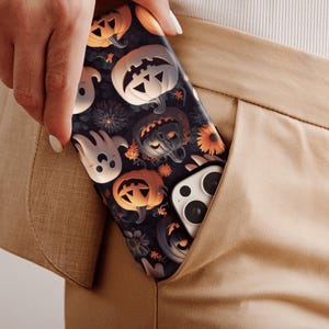 May include: A black phone case with a Halloween theme. The case features a pattern of pumpkins, ghosts, and flowers. The phone case is in a pocket of a pair of brown pants.