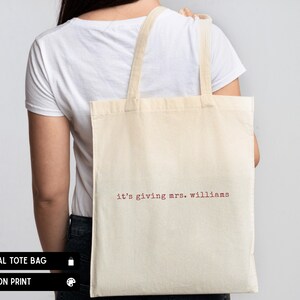 K&ouml;nnte beinhalten: Eine naturfarbene Stofftasche mit dem Text "it's giving mrs. williams", der in einer karmesinroten Schreibmaschinenschrift aufgedruckt ist. Die Tasche hat zwei Schulterriemen und eine rechteckige Form f&uuml;r ein schlichtes und funktionales Design.