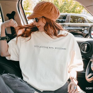 K&ouml;nnte beinhalten: Ein elfenbeinfarbenes T-Shirt mit dem schwarzen Schriftzug 'it's giving mrs. williams' auf der Brust. Das T-Shirt hat einen Rundhalsausschnitt und kurze &Auml;rmel, was f&uuml;r eine lockere Passform sorgt. Der Stoff ist ein einfarbiges Material.