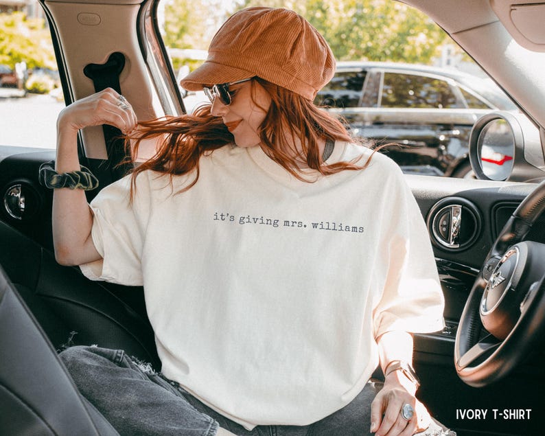 K&ouml;nnte beinhalten: Ein elfenbeinfarbenes T-Shirt mit dem schwarzen Schriftzug 'it's giving mrs. williams' auf der Brust. Das T-Shirt hat einen Rundhalsausschnitt und kurze &Auml;rmel, was f&uuml;r eine lockere Passform sorgt. Der Stoff ist ein einfarbiges Material.