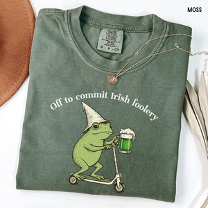 May include: Moss green t-shirt with the phrase "Off to commit Irish foolery" and a cartoon frog wearing a hat and holding a beer mug while riding a scooter. A silver necklace with a pendant is also visible.