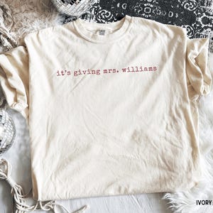 K&ouml;nnte beinhalten: Ein elfenbeinfarbenes Kurzarm-T-Shirt mit dem Text 'it's giving mrs. williams', der in einer dunkelroten, kleingeschriebenen Schriftart auf der Brust aufgedruckt ist. Das Kleidungsst&uuml;ck hat einen Rundhalsausschnitt und ein sichtbares Markenetikett.