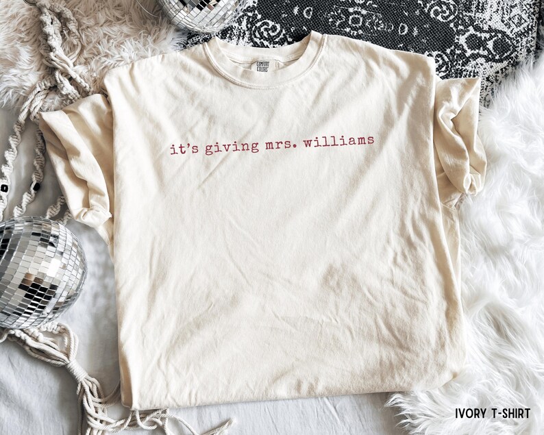 K&ouml;nnte beinhalten: Ein elfenbeinfarbenes Kurzarm-T-Shirt mit dem Text 'it's giving mrs. williams', der in einer dunkelroten, kleingeschriebenen Schriftart auf der Brust aufgedruckt ist. Das Kleidungsst&uuml;ck hat einen Rundhalsausschnitt und ein sichtbares Markenetikett.