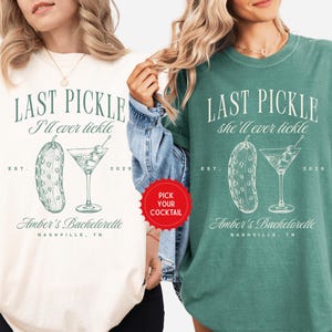 May include: Two t-shirts, one cream and one green, with a graphic design. The design includes the text "LAST PICKLE I'll ever tickle" and "LAST PICKLE she'll ever tickle", a pickle illustration, and a martini glass. The text "Amber's Bachelorette Nashville, TN" is also present.