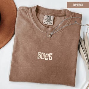 May include: A brown t-shirt with the number "8647" printed on the front in white. The shirt is made of a soft, comfortable fabric and has a distressed look.