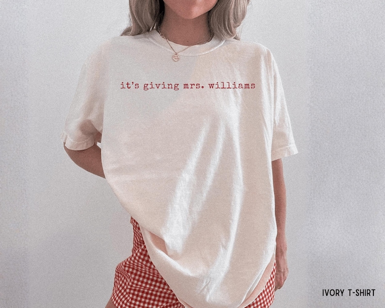 K&ouml;nnte beinhalten: Ein elfenbeinfarbenes Kurzarm-T-Shirt mit dem roten Schriftzug "it's giving mrs. williams" auf der Brust. Das Kleidungsst&uuml;ck hat einen Rundhalsausschnitt und eine lockere, weit geschnittene Passform, kombiniert mit einer rot-wei&szlig; karierten Shorts.