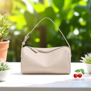 Puede incluir: Un bolso de mano de color beige claro con forma de baguette, con cierre de cremallera en la parte superior y un solo asa superior. El bolso está sobre una superficie blanca con un fondo verde frondoso.