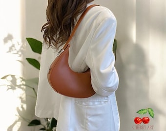 May include: A brown leather half-moon shaped handbag with a silver clasp. The bag is worn by a person wearing a white blazer.