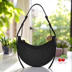 May include: A black crescent-shaped handbag with a long, adjustable strap. The bag has a smooth, matte finish and a curved design. The background features greenery and a white table, suggesting an outdoor setting. The bag is a stylish accessory.