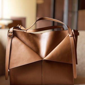 May include: A brown leather tote bag with a unique geometric design. The bag features a structured shape with folded panels and long straps for carrying. The bag has a buckle closure and is made of smooth, high-quality leather.