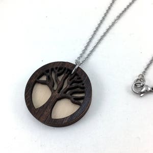 May include: A dark brown wooden necklace with a tree of life pendant. The pendant is a circle with a cutout of a tree with branches and leaves. The necklace is on a silver chain.
