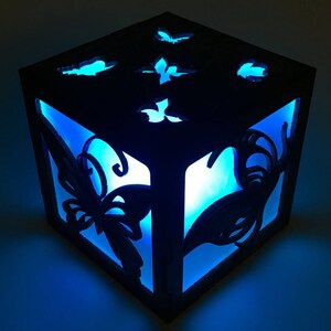 May include: A dark wooden cube-shaped lamp with butterfly cutouts and a bright blue interior glow. The butterfly designs are visible on the top and sides of the lamp. The lamp is illuminated, creating a soft, ambient light.
