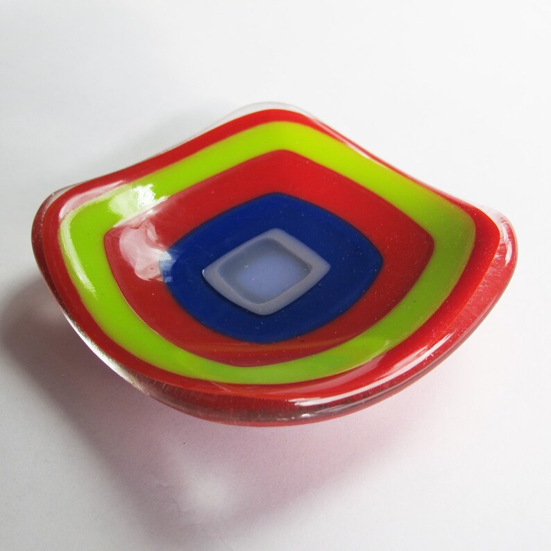 May include: A small, red glass dish with concentric circles of yellow, blue, and white. The dish is slightly curved and has a glossy finish.