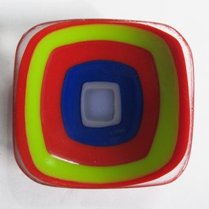 May include: A square glass dish with concentric circles of red, yellow and blue glass.