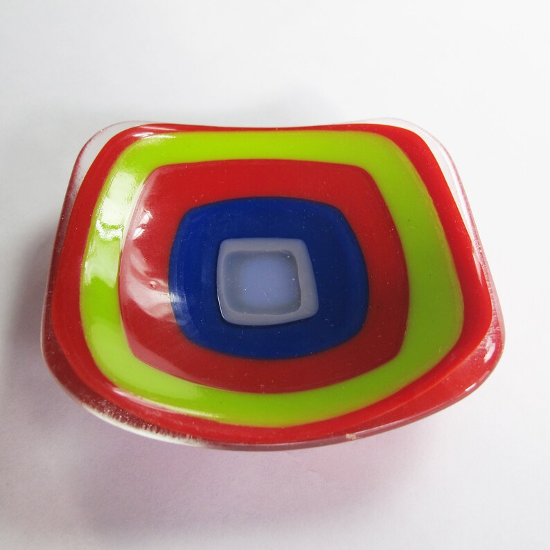 May include: A small, red, yellow and blue glass dish with a concentric circle design. The dish is made of fused glass and has a slightly rounded shape.
