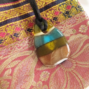 May include: A handcrafted pendant necklace with a distinctive, layered design. The pendant showcases a blend of colours including teal, brown, yellow, and white, set in a clear, glossy resin. The necklace has a black cord.