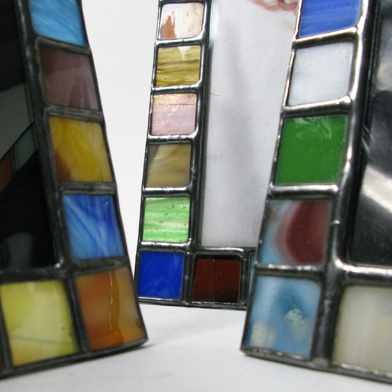 Op de afbeelding: Close-up van drie glas-in-lood fotolijsten. Elke lijst heeft een geometrisch ontwerp met kleurrijke glazen vierkanten in tinten blauw, groen, geel en rood, gezet in een metalen frame. De lijsten worden op een wit oppervlak weergegeven.