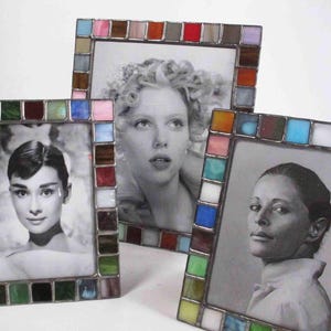 Op de afbeelding: Drie vierkante fotolijsten met kleurrijke glas-in-loodranden. De lijsten bevatten zwart-wit portretten van vrouwen. De glazen stukken zijn in verschillende kleuren, waaronder groen, blauw, roze en bruin. De lijsten zijn op een wit oppervlak geplaatst.