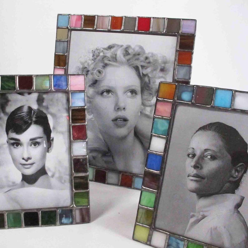 Op de afbeelding: Drie vierkante fotolijsten met kleurrijke glas-in-loodranden. De lijsten bevatten zwart-wit portretten van vrouwen. De glazen stukken zijn in verschillende kleuren, waaronder groen, blauw, roze en bruin. De lijsten zijn op een wit oppervlak geplaatst.