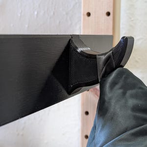 May include: A black, 3D-printed coat hook attached to a wooden frame. The hook has a curved design and is mounted on a black rectangular base. The coat hook is designed to hold a coat.