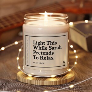 May include: A lit candle in a glass jar with a label that reads "Light This While Sarah Pretends To Relax." The candle sits on a round wooden coaster, surrounded by string lights. The background includes a guitar and a book.