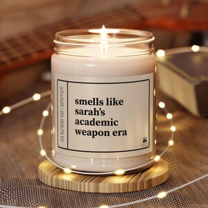 May include: A lit candle in a glass jar with the text "smells like sarah's academic weapon era" on a white label. The candle sits on a round wooden coaster, surrounded by string lights. The background includes a guitar and a book.