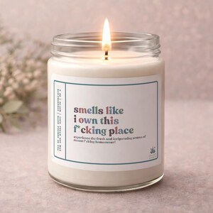 May include: A lit, glass jar candle with a label that reads "smells like I own this f*cking place." The candle has a white wax and a blue and white label. The text on the label also says "experience the fresh and invigorating aroma of recent f*cking homeowner!"
