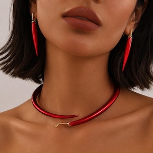 Może przedstawiać: Zestaw biżuterii składający się z naszyjnika typu choker w kształcie czerwonej papryczki chili oraz pasujących kolczyków. Naszyjnik ma zakrzywiony, błyszczący czerwony wzór papryczki ze złotą łodyżką, a kolczyki to długie, czerwone szpiczaste elementy.