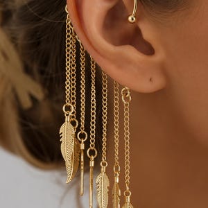 May include: A gold-toned ear cuff featuring a curved wire that hooks over the upper ear, with multiple dangling chains of varying lengths. Several small, textured metal feather charms and thin, straight metal bars are attached to the chains.