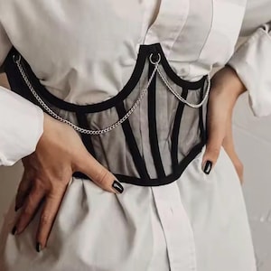 May include: A black corset-style waist belt featuring sheer mesh panels, structured black boning, and a silver-toned metal chain draped across the front. The garment is worn over a crisp white button-down shirt with long sleeves and cuffed wrists.