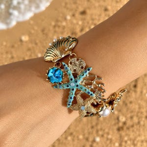 May include: A gold-toned charm bracelet featuring various nautical elements including a blue starfish with small crystals, a textured seashell, a sand dollar, a coral-shaped piece, and a white pearl bead, all linked together in a cohesive design.